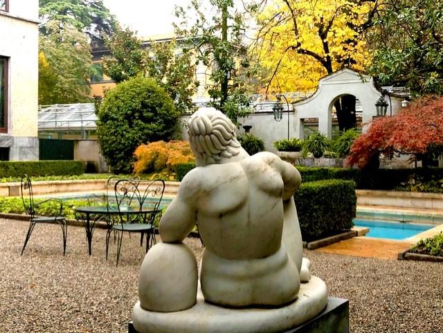 Rear view of a classical marble statue seated in a quiet garden courtyard, surrounded by manicured hedges, autumn-colored trees, a small pool of water, and wrought-iron café chairs near a pale stucco building.