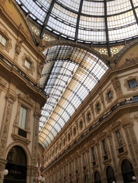 Interior view of an elegant historic shopping arcade with ornate cream-colored façades, arched windows, and decorative moldings, topped by a sweeping glass-and-iron vaulted ceiling that fills the space with natural light.