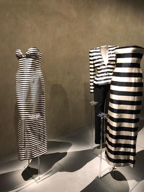 Two black-and-white striped garments displayed on minimalist mannequins in a museum or gallery setting, lit dramatically against a neutral wall, emphasizing bold horizontal stripes and sculptural silhouettes.