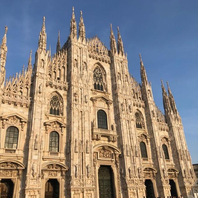 The Duomo di Milano’s ornate Gothic façade, with towering spires, intricate stone carvings, and arched windows, illuminated by daylight against a clear blue sky.