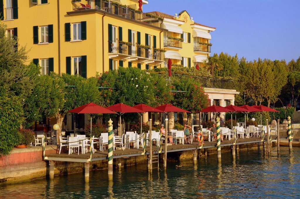 Outdoor waterfront café with red umbrellas and white seating along a canal, in front of a yellow building with balconies and green shutters on a sunny day.