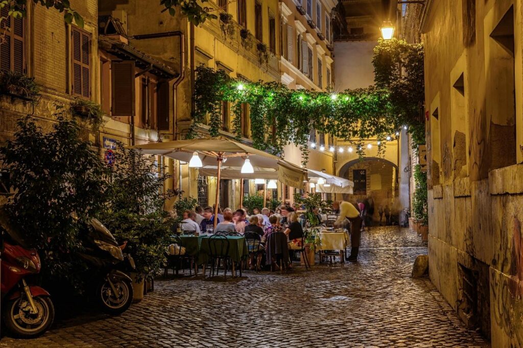 A cozy Rome side street at night with outdoor café tables filled with diners, warm string lights overhead, ivy-covered walls, and a narrow cobblestone lane creating an intimate, atmospheric scene.
