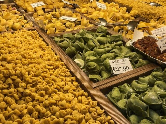 Fresh handmade pasta displayed at an Italian market, with trays of yellow and green tortellini and balanzoni neatly arranged and labeled with prices.
