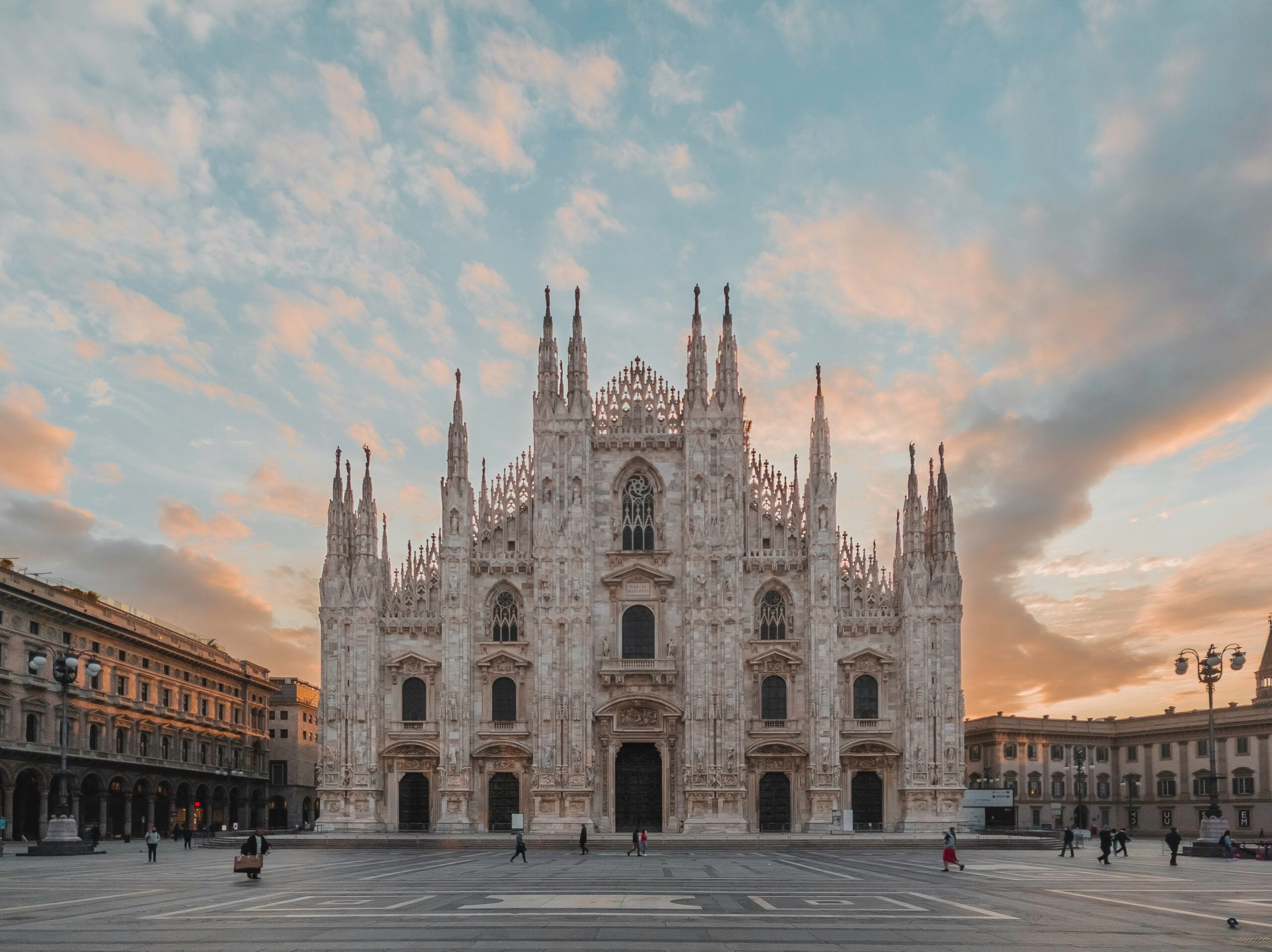 Front view of the Duomo di Milano (Milan Cathedral) in Piazza del Duomo at sunset, with a pastel pink and blue sky and a few people walking across the wide stone square.