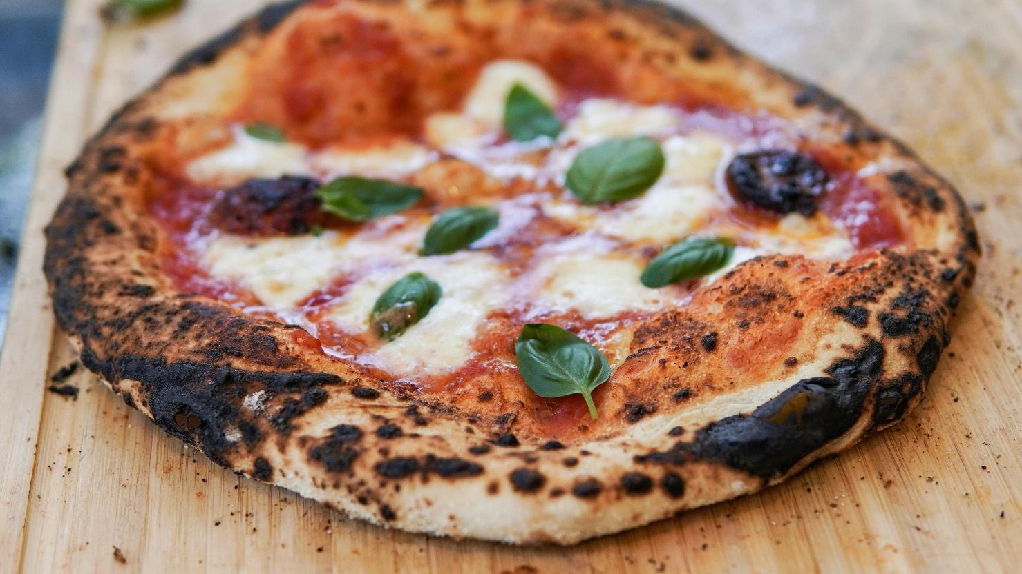 A Neapolitan-style margherita pizza with a charred, blistered crust topped with San Marzano tomato sauce, fresh mozzarella, and whole basil leaves, resting on a wooden pizza board — the best pizza in Naples.