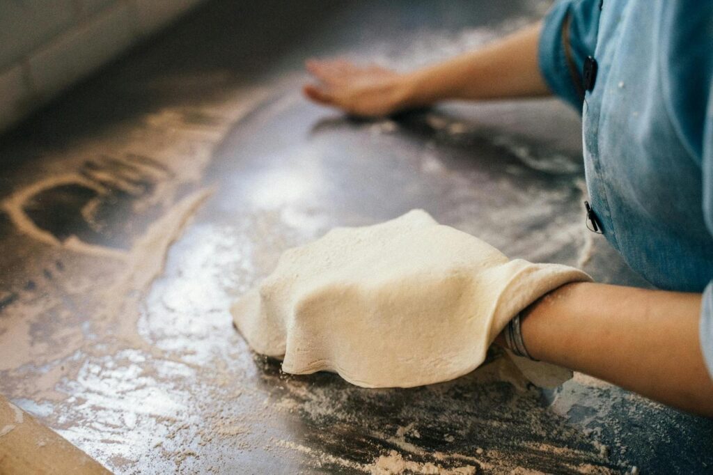 A pizzaiolo stretching fresh pizza dough by hand on a flour-dusted dark metal work surface, wearing a blue apron in a professional pizzeria kitchen.