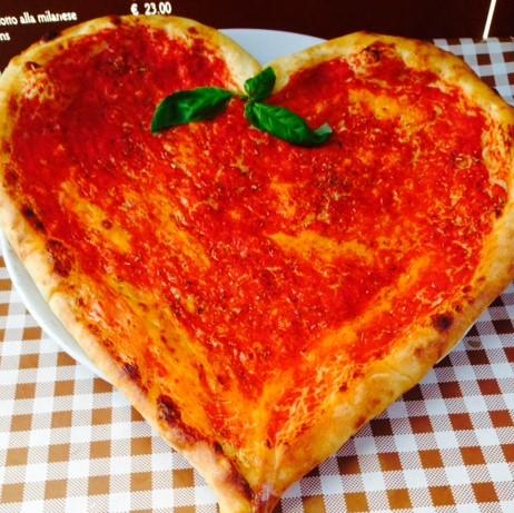 A heart-shaped margherita pizza topped with vibrant tomato sauce and two fresh basil leaves, served on a white plate over a classic red-and-white checkered tablecloth at an Italian restaurant.