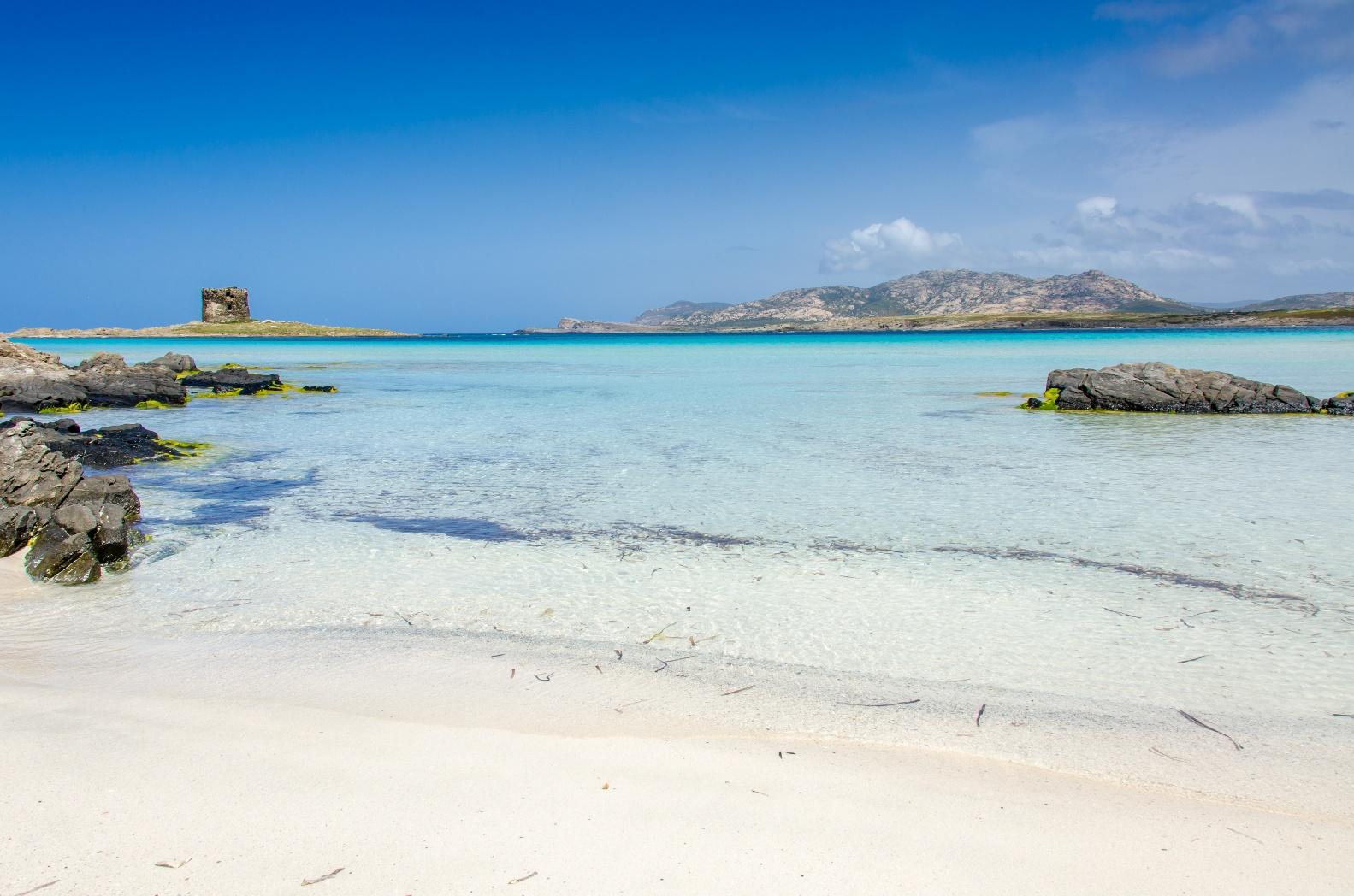 Crystal-clear turquoise water and white sand beach on the northern coast of Sardinia with rocky shoreline and a historic coastal tower in the distance.