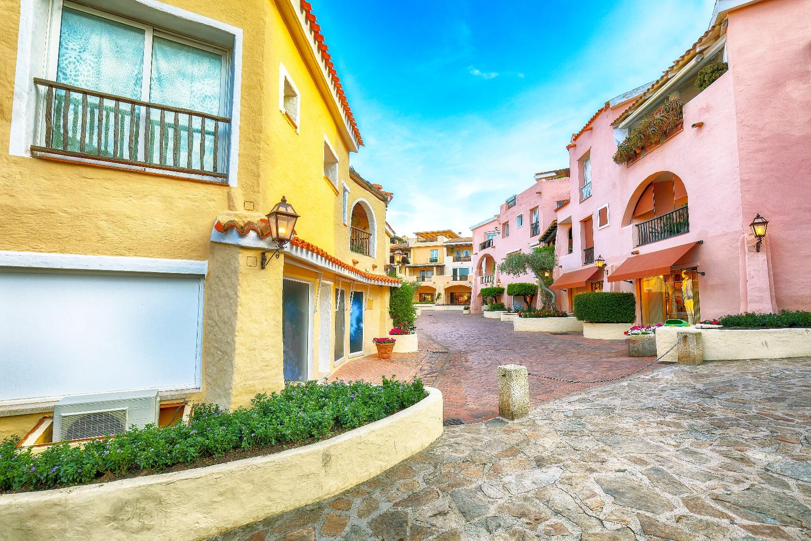 Colorful pastel buildings lining a charming cobblestone street in a seaside village in Sardinia, featuring balconies, shutters, and Mediterranean architecture.