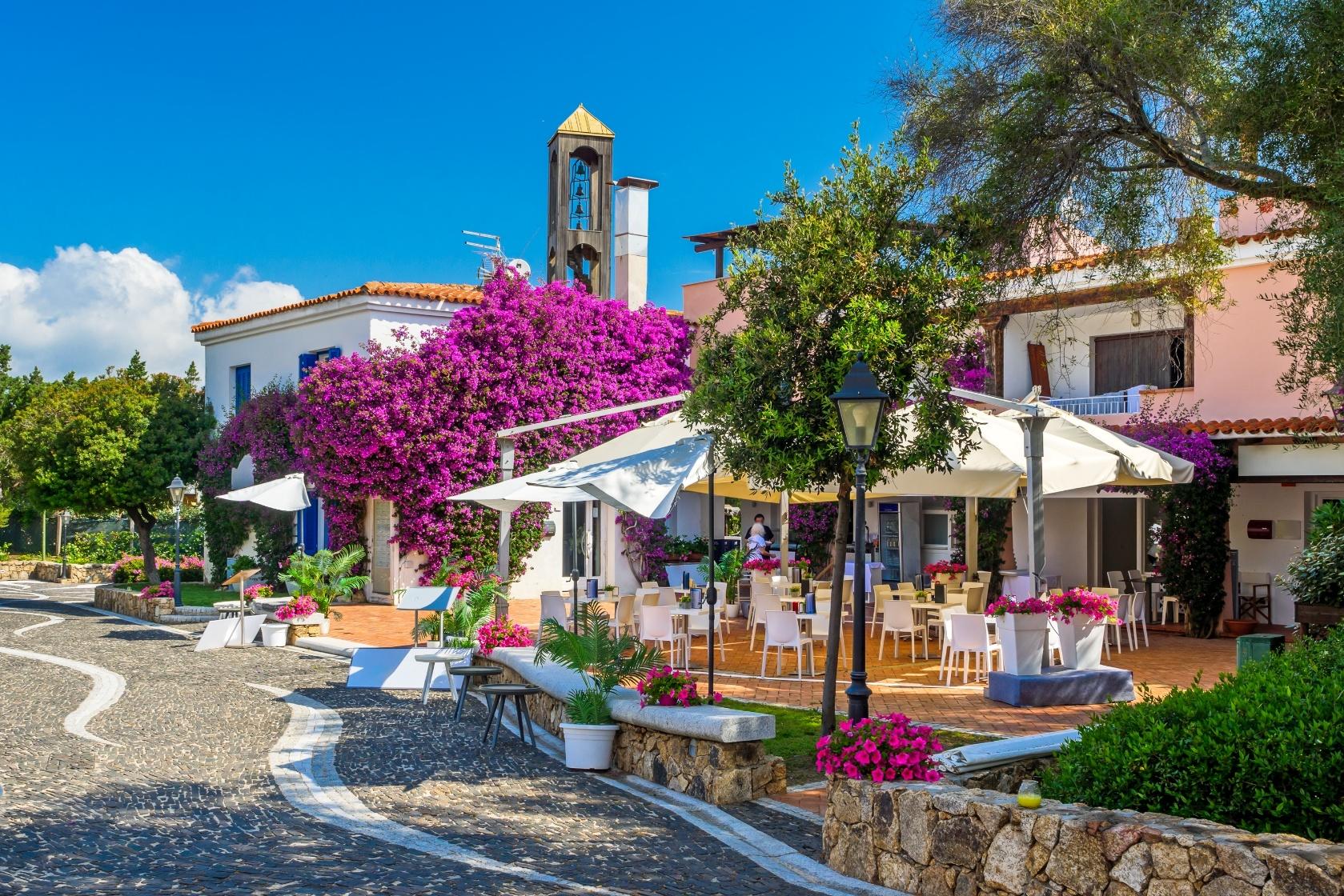 Scenic outdoor café with blooming bougainvillea and white umbrellas in a picturesque village in Sardinia, set along a quiet cobblestone road under a bright blue sky.