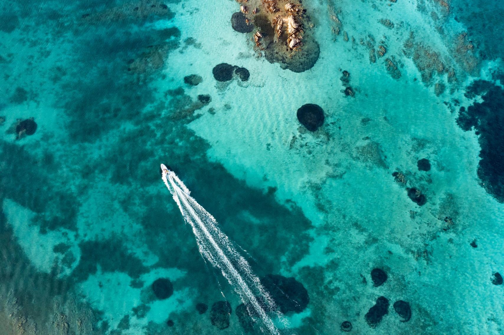 Aerial view of a small motorboat speeding through clear turquoise waters over rocky reefs along Sardinia’s coastline.