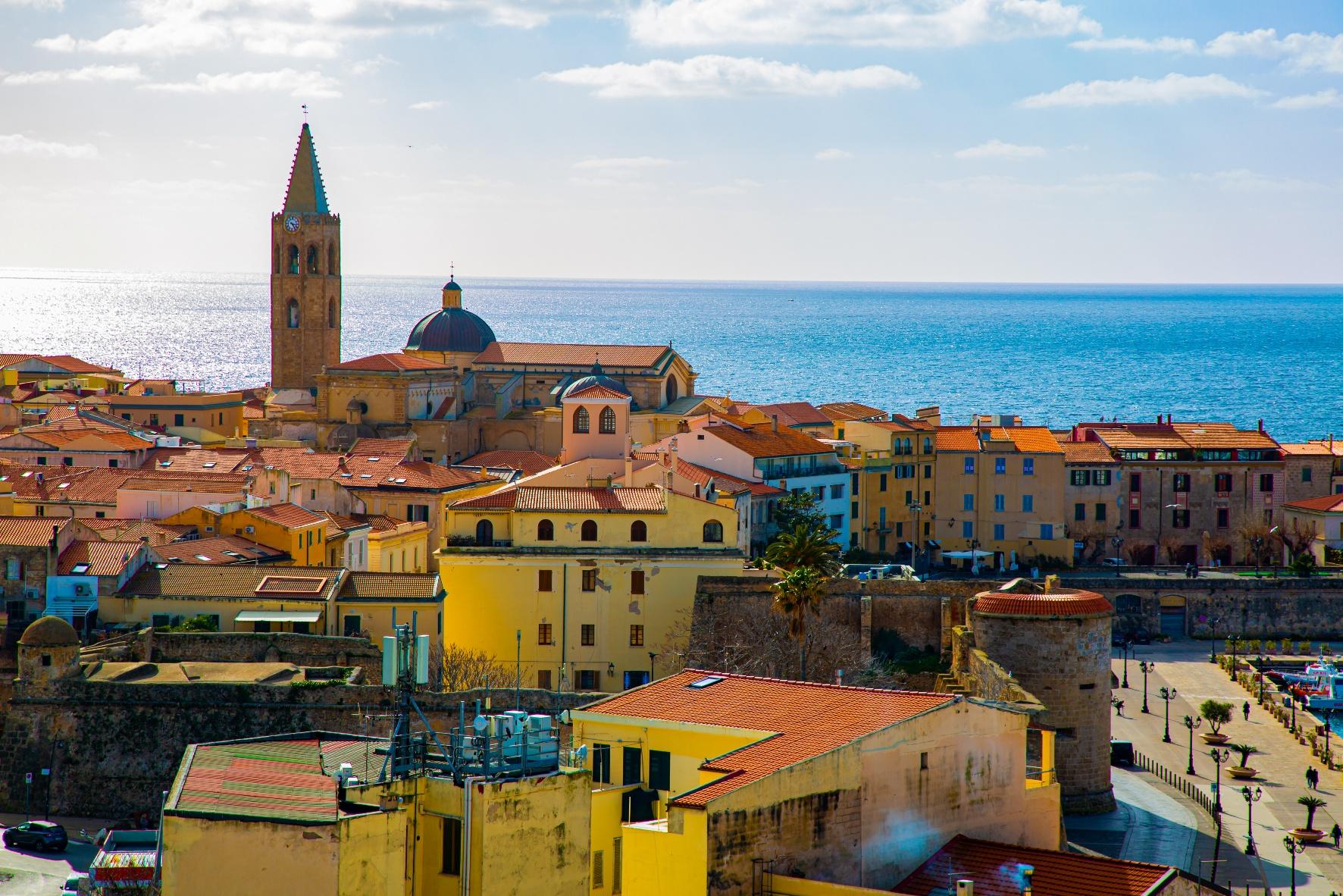 Colorful seaside town in Sardinia with terracotta rooftops, a historic church tower, and the Mediterranean Sea in the background.