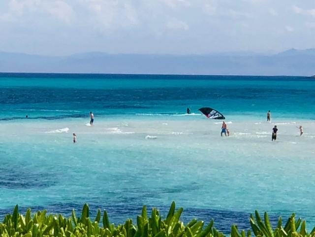 People wading in shallow, crystal-clear water on a Sardinian beach with shades of blue sea and distant mountains on the horizon.
